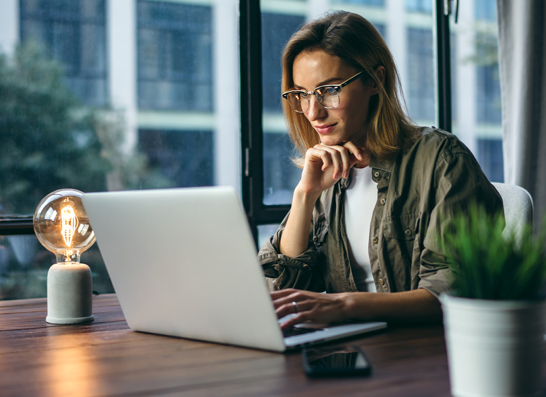 Video Library - Woman Curiously Looking at Open Laptop While Sitting at a Wooden Table with a Light Bulb